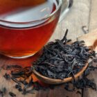 Tasse en verre de thé noir Assam rouge-ambré avec grandes feuilles noires sur cuillère en bois fond rustique